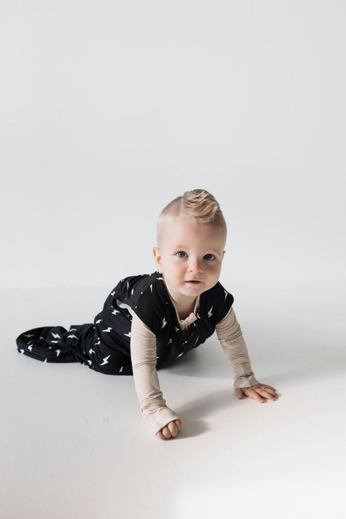 A baby wearing the 'forever french baby' Bamboo Sleep Sack in the Midnight Lightning Bolt design over a long-sleeve beige shirt is crawling on a white surface. The baby, with light-colored hair and blue eyes, looks directly at the camera with a curious expression. This luxurious sleep outfit ensures comfort throughout the night.