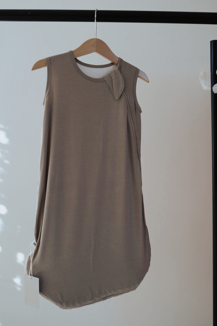The Bamboo Sleep Sack in grey by forever french baby hangs on a wooden hanger against a plain backdrop, showing off its sleeveless design, round neckline, unique fabric detail at the shoulder, and a white tag at the bottom left.