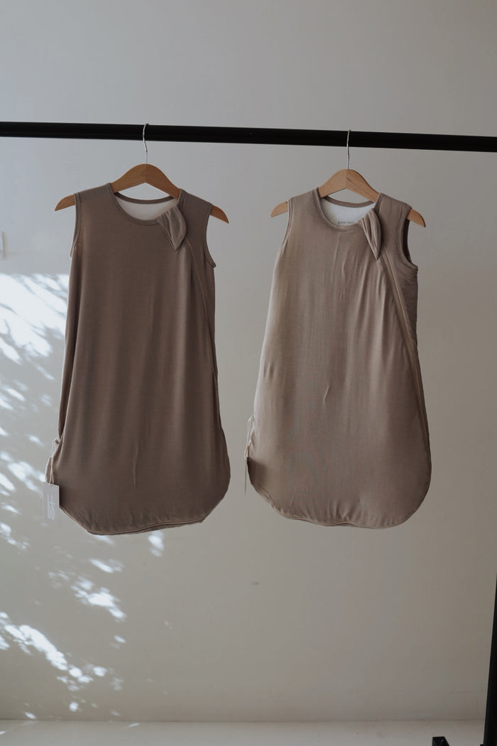 Two grey Bamboo Sleep Sacks from forever french baby with shoulder snaps hang side by side on wooden hangers from a black rod against a light wall, dappled in sunlight—perfect as cozy wearable blankets for your baby.