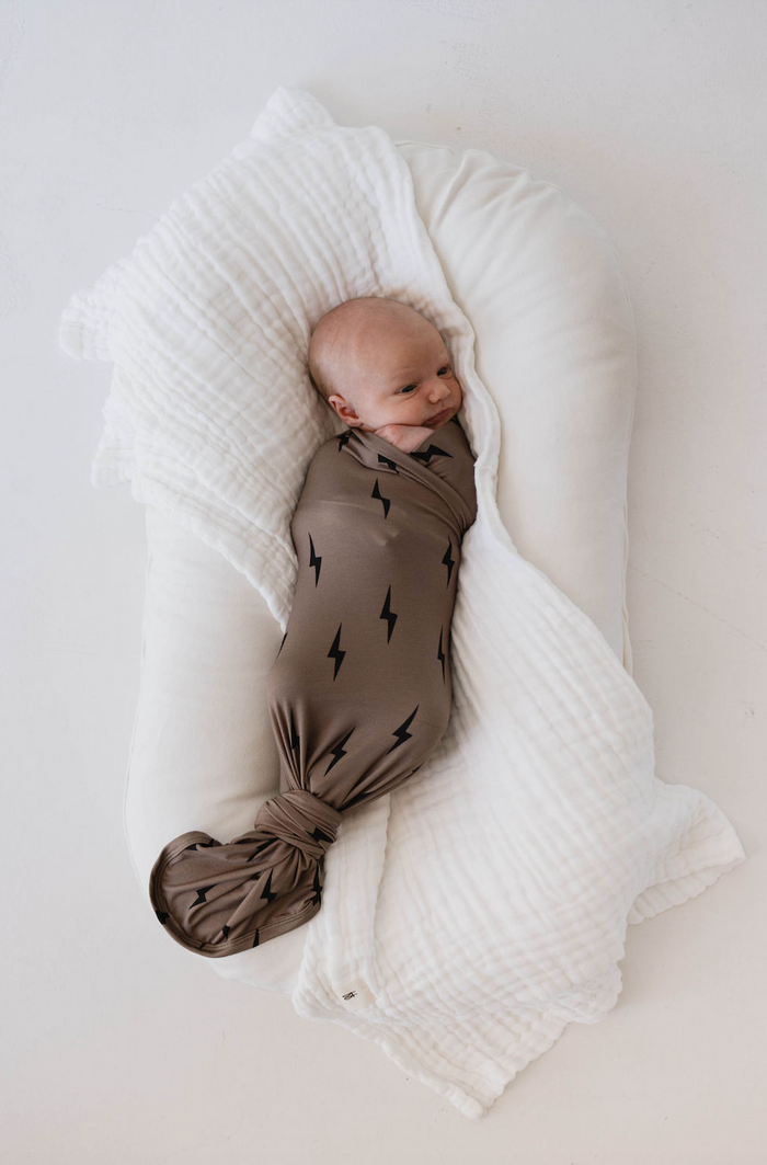 A newborn baby is wrapped snugly in the "Bamboo Swaddle | Black & Brown Lightning Bolt" from forever french baby, lying on a white cushion with a textured white blanket. The breathable, hypo-allergenic material ensures the baby is facing slightly upward and appears calm and content.