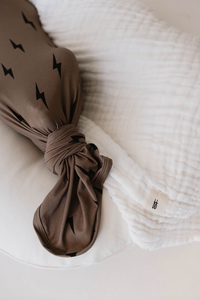 A Bamboo Swaddle from forever french baby featuring black and brown lightning bolt patterns is tied in a knot and placed on quilted white fabric. The hypo-allergenic swaddle has a small tag with the initials "bf" visible on the right side of the image.