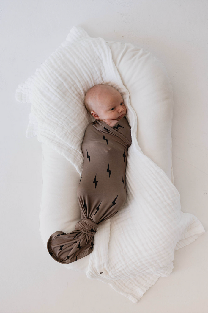A newborn baby is swaddled in a forever french baby Bamboo Swaddle | Black & Brown Lightning Bolt blanket, lying on a large white cushion. The baby appears calm and content against the plain white background.