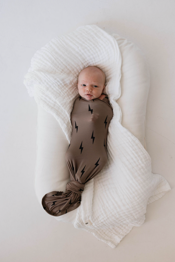 A baby is swaddled in the forever french baby Bamboo Swaddle | Black & Brown Lightning Bolt, lying on a cushioned white surface with a hypo-allergenic plush blanket partially covering them. The baby has a calm expression and is looking upwards.