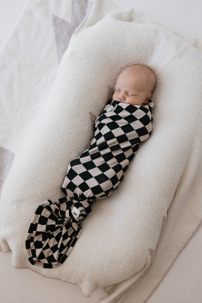 A baby is peacefully sleeping, swaddled in the Bamboo Swaddle | Black Checkerboard from forever french baby. The baby is lying on a soft, white, textured cushion. The background is light and neutral, enhancing the calm and serene atmosphere.
