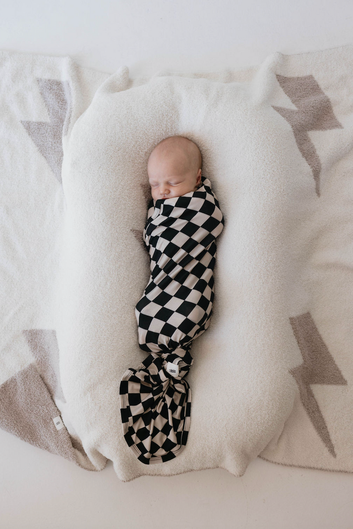 A baby wrapped in a Black Checkerboard Bamboo Swaddle from forever french baby is sleeping on a white plush blanket with light grey patterns on either side, lying peacefully in the center.