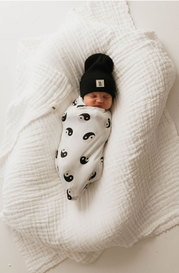 A sleeping baby swaddled in a Bamboo Swaddle | Black & White Yin Yang by forever french baby rests on a crumpled white quilt. The baby is wearing a black knit hat, and the background is plain white.