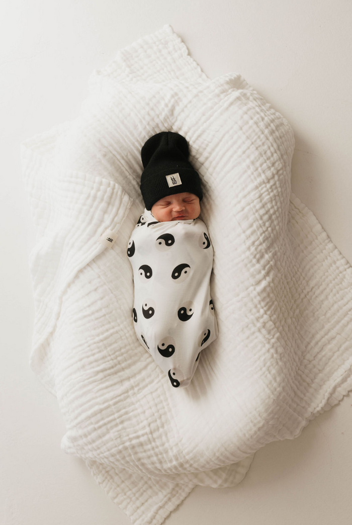 A newborn baby is enveloped in a hypo-allergenic, breathable blanket named Bamboo Swaddle | Black & White Yin Yang by forever french baby, featuring black yin-yang symbols. The baby is also sporting a black beanie hat and is resting on a white, textured, crumpled blanket. The baby's eyes are closed, and they appear to be peacefully sleeping.