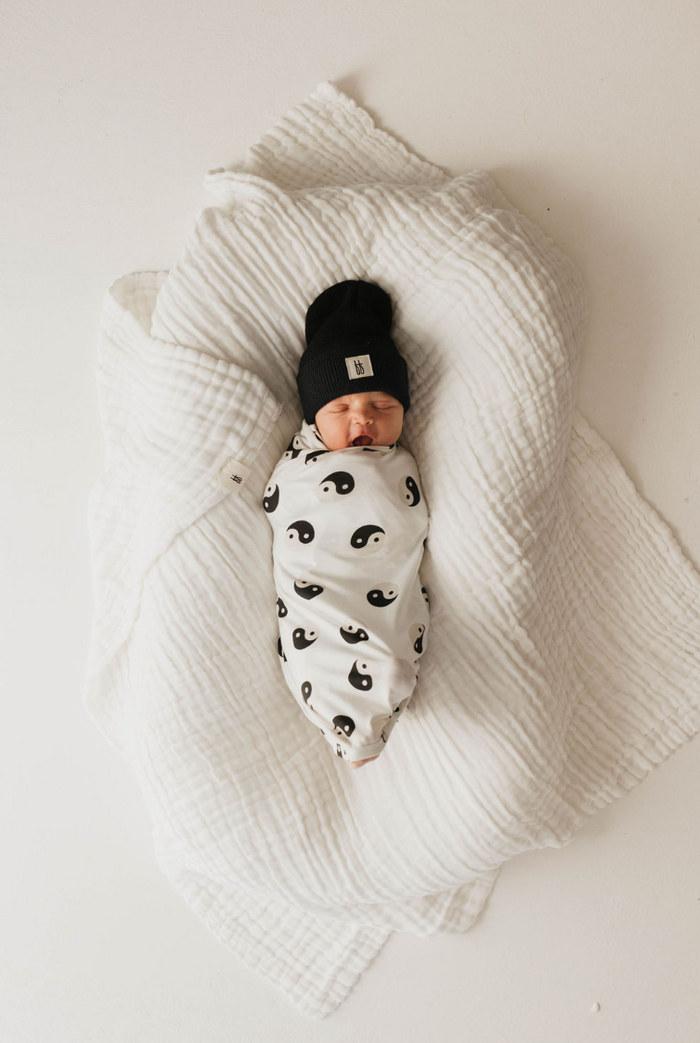 A newborn baby is swaddled in a hypo-allergenic forever french baby Bamboo Swaddle | Black & White Yin Yang. The baby lies on a large white quilt, wearing a black knit hat, against the plain white background.