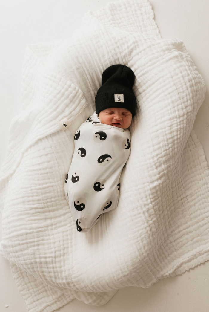 A newborn baby lies peacefully sleeping on a soft, white, textured blanket, swaddled in the "Bamboo Swaddle | Black & White Yin Yang" by forever french baby and wearing a black beanie.