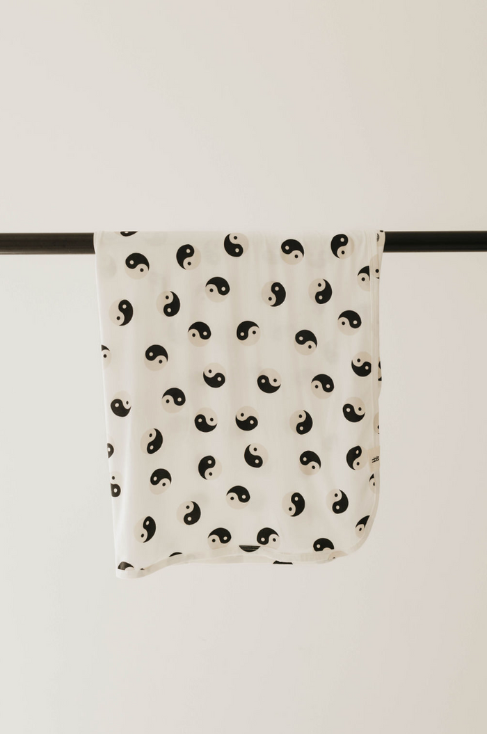 A Bamboo Swaddle with a black and white yin-yang pattern from Forever French Baby, made from breathable fabric, is draped over a black horizontal rod against a plain beige background.