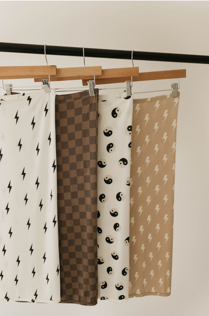 Four patterned fabrics hang on a wooden rod with metal clips. The breathable swaddles, from left to right, feature black lightning bolts on white, a brown checkered design, the Bamboo Swaddle | Black & White Yin Yang by forever french baby, and white lightning bolts on beige.