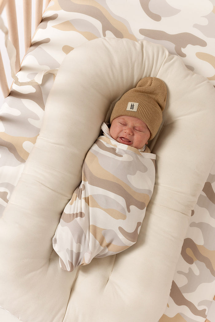 A newborn baby in the forever french baby Bamboo Swaddle | Camo 2.0 and a brown knit beanie rests in a soft cream lounger against a matching camouflage-patterned background.
