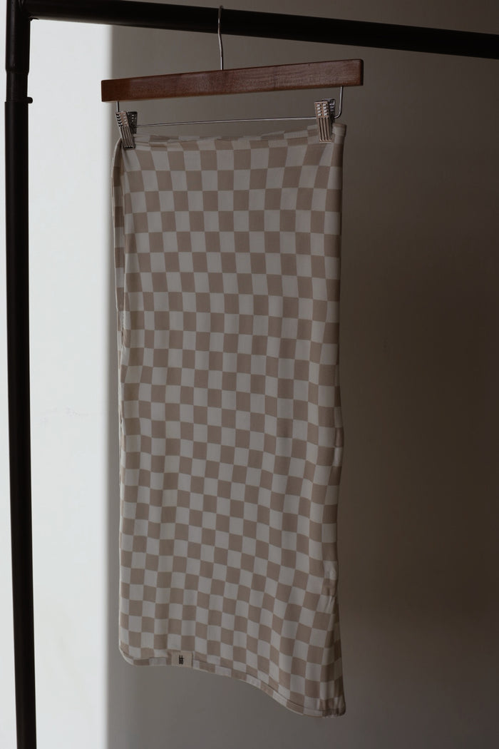 The forever french baby Bamboo Swaddle | Checkerboard, in beige and white checkered fabric, hangs from a wooden hanger on a black clothing rack against a softly lit plain background, echoing gentle comfort and breathability.