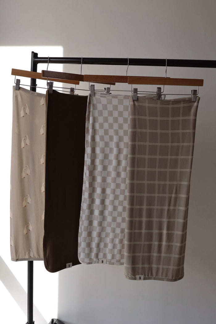 Four towels from forever french baby, including the Bamboo Swaddle | Checkerboard, hang on a black rack. The designs feature abstract shapes, brown, checkered, and grid patterns in neutral tones as sunlight filters in.