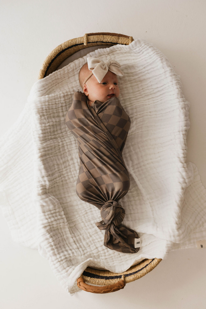 A baby, swaddled in a "Faded Brown Checkerboard" Bamboo Swaddle from Forever French Baby and wearing a beige headband with a bow, lies in a wicker basket on a soft white blanket. The baby is gazing to the side, creating a peaceful and cozy scene.