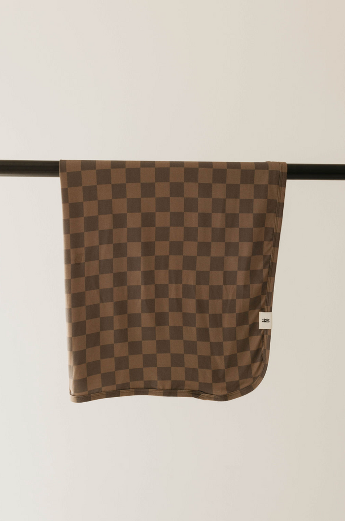 A Bamboo Swaddle in Faded Brown Checkerboard from forever french baby, perfect for cozy evenings, hangs neatly folded over a horizontal black rod against a plain off-white background. A small white tag is visible on the swaddle, subtly hinting at its hypo-allergenic qualities.