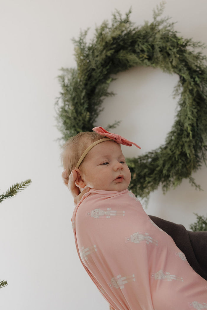 A baby in the forever french baby Bamboo Swaddle | Nutcracker is lifted up, wearing a pink headband. A green wreath decorates the white wall in the background.