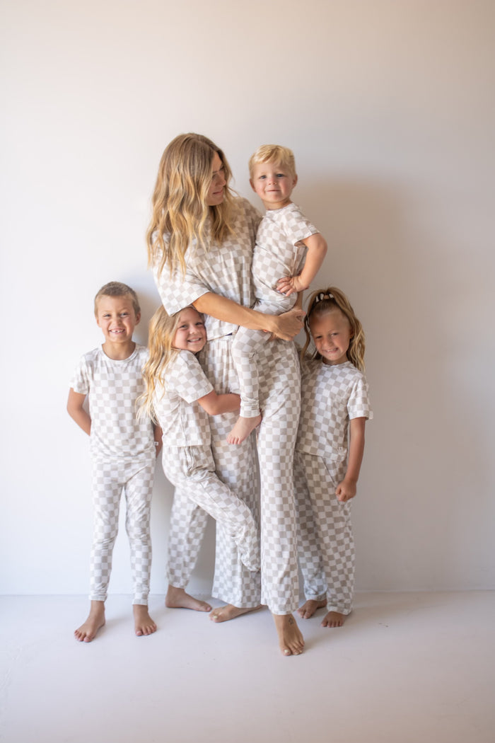 A woman and four young children smile together in matching forever french baby Bamboo Two Piece Lounge Set | Checkerboard pajamas, standing before a plain off-white wall.