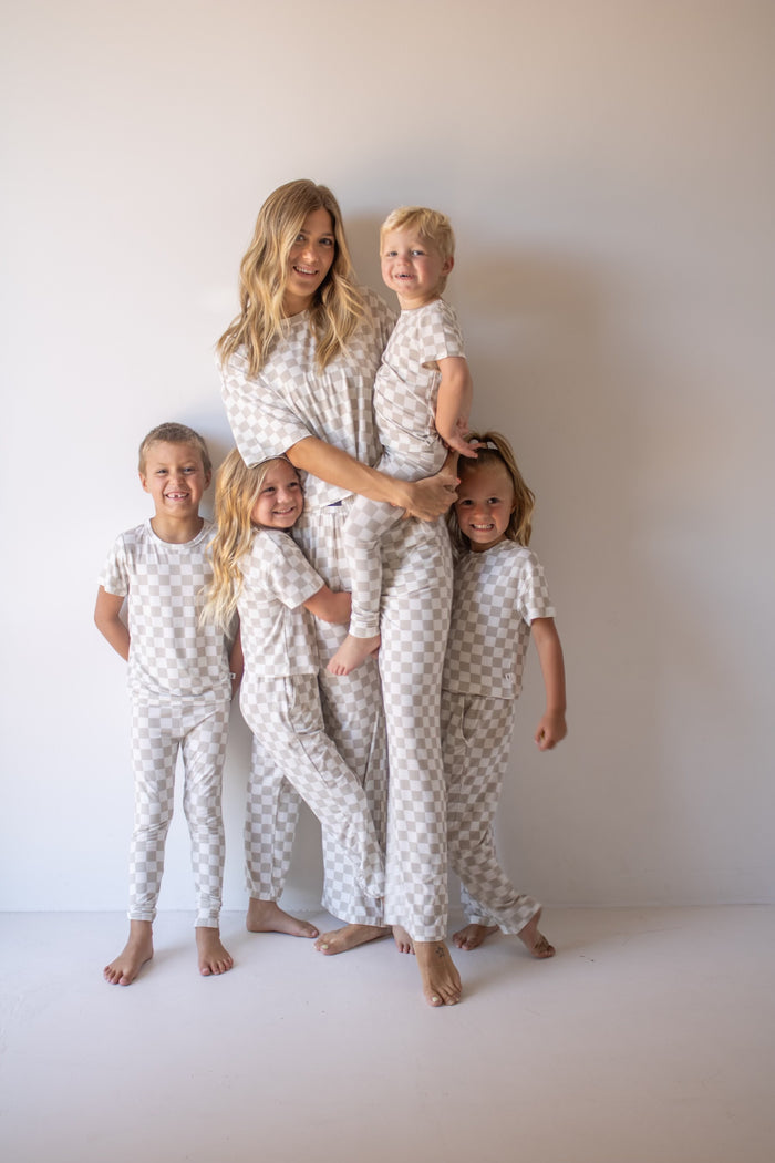 A woman and four children smile barefoot against a light wall, all wearing forever french baby Women's Boxy Tee Lounge Sets in a light checkerboard pattern, radiating cozy family loungewear vibes.