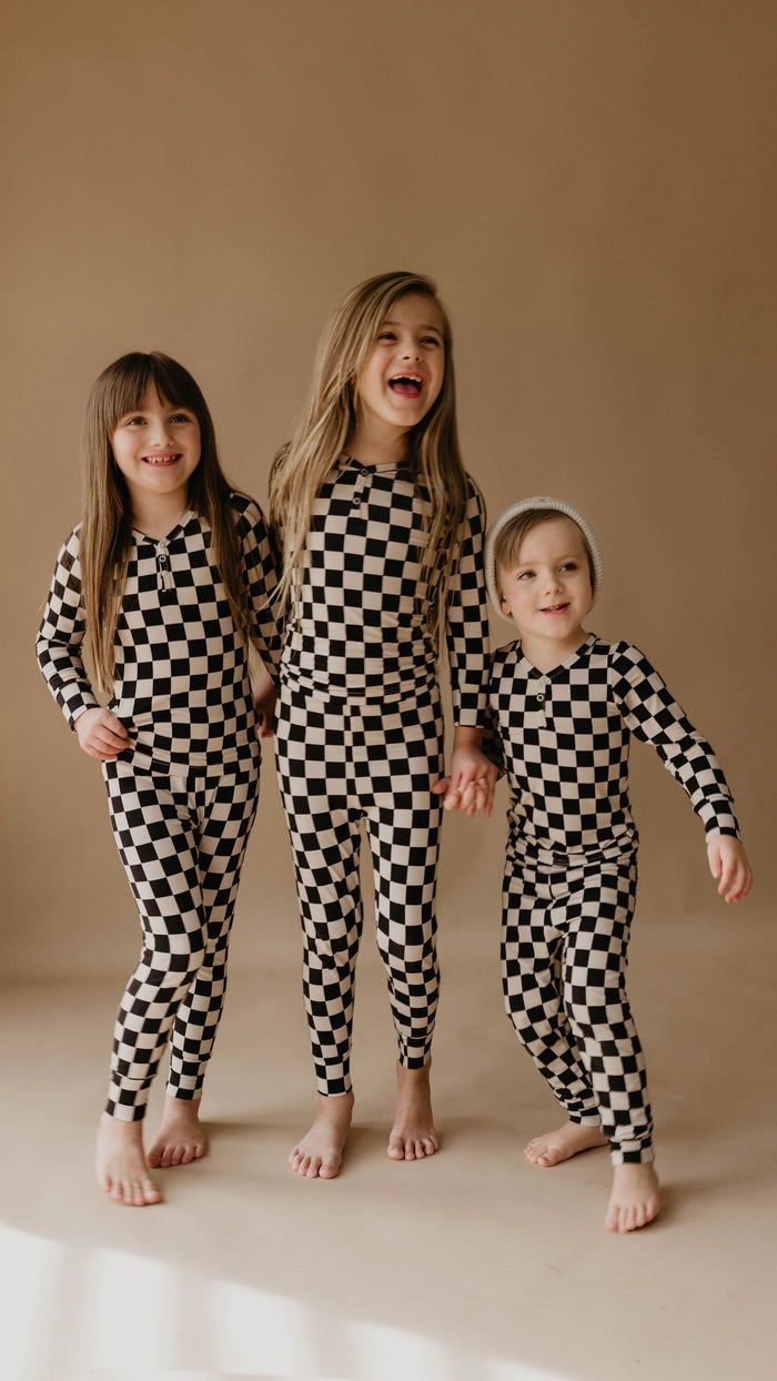 Three children are smiling and standing in a row against a plain background, all wearing matching Bamboo Two Piece Pajamas in Black Checkerboard from forever french baby. The child in the middle has their mouth open in a laugh, while the child on the right sports a knit hat. They are holding hands.