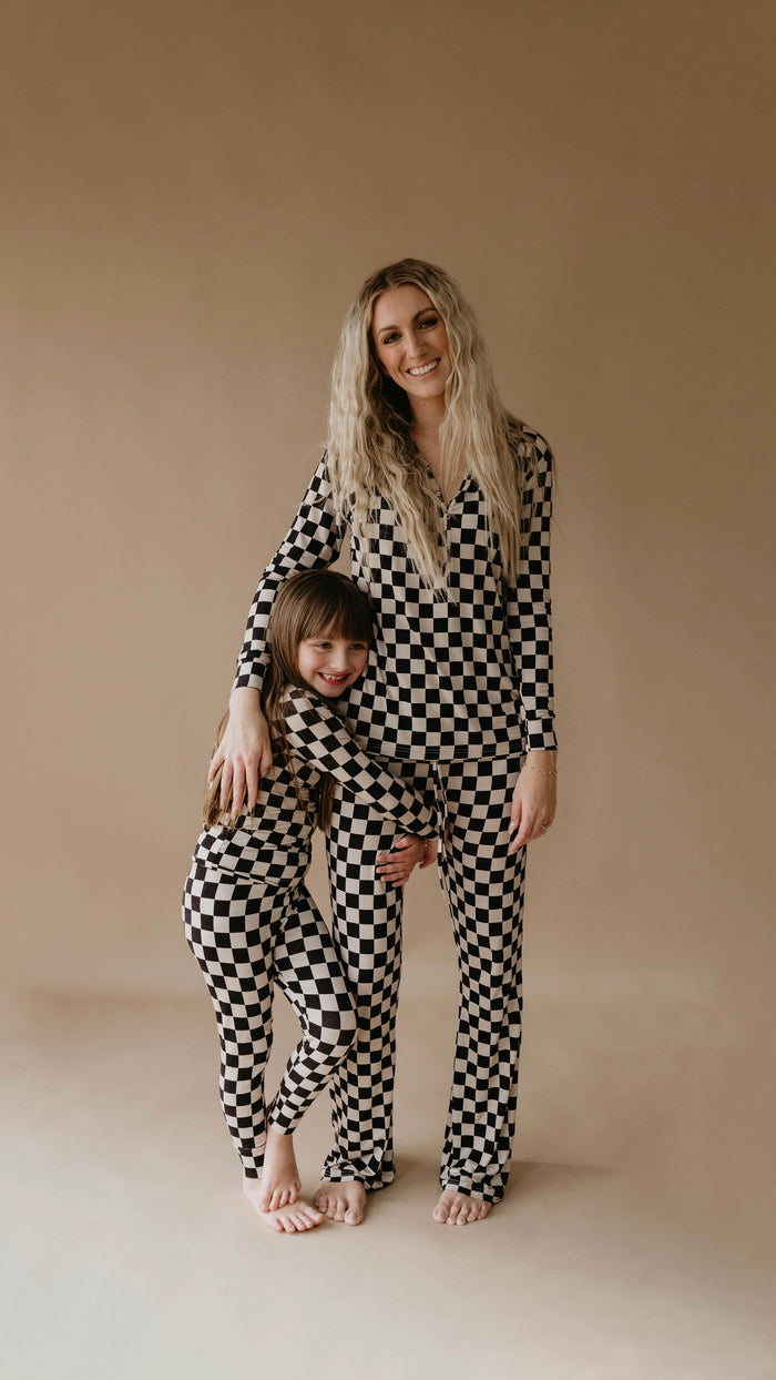 A woman and a child stand together, both wearing matching Bamboo Two Piece Pajamas in Black Checkerboard from forever french baby. The child hugs the woman's waist, and both have bare feet. The background is a simple, light brown color. They both look happy and relaxed.