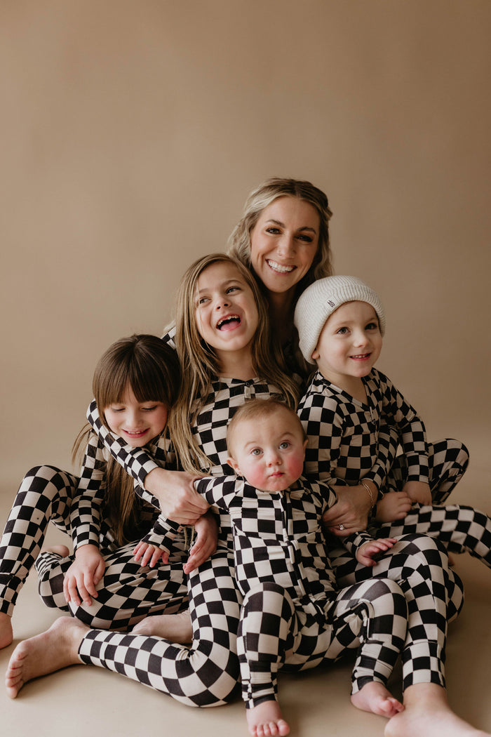 A smiling woman sits hugging four young children, all dressed in forever french baby Bamboo Two Piece Pajamas in a black checkerboard design, against a plain beige background. One of the children wears a white beanie.