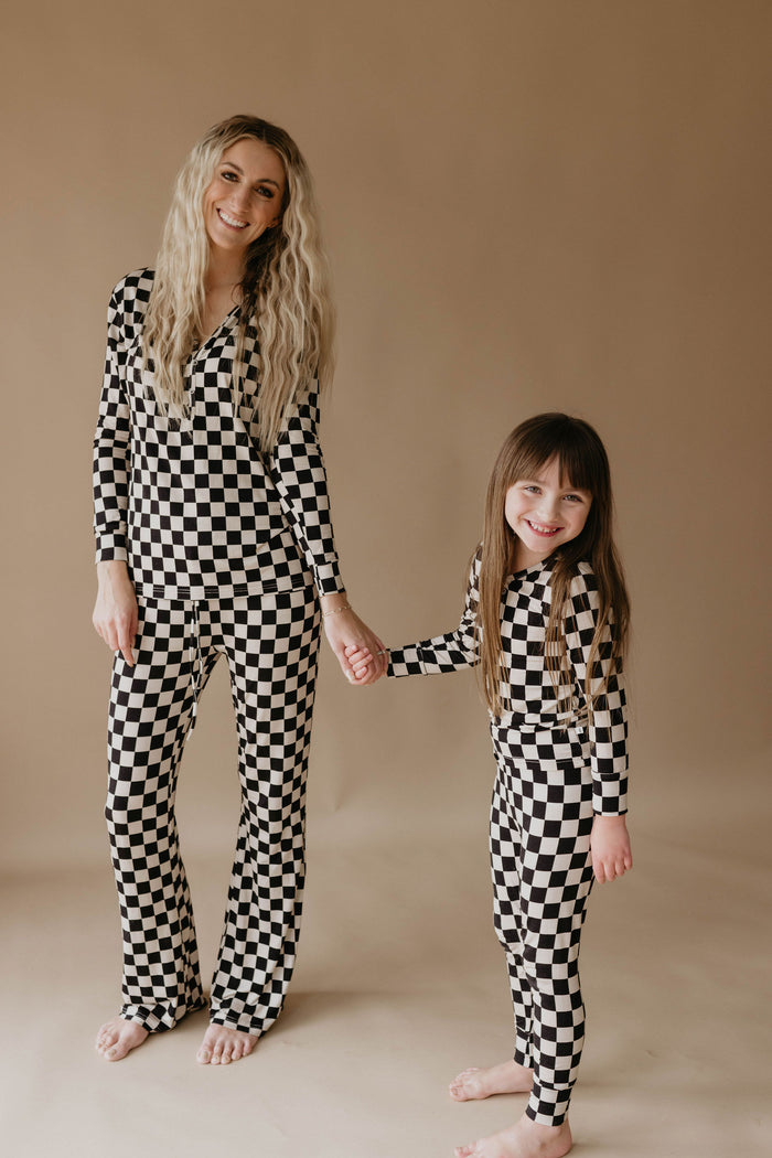 A woman and a young girl stand holding hands and smiling at the camera, both wearing matching Forever French Baby Bamboo Two Piece Pajamas in Black Checkerboard. The plain beige background creates a warm atmosphere perfect for their Forever French Baby moments.