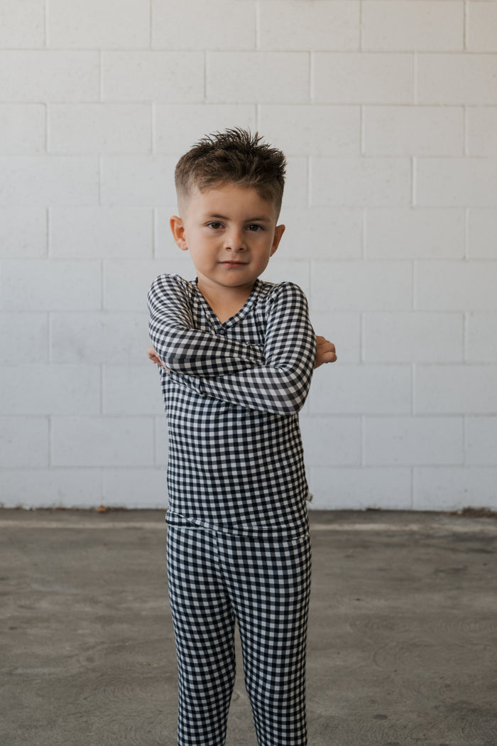 A young child with short hair stands confidently on a concrete floor, arms folded, wearing the forever french baby Bamboo Two Piece Pajamas in Black Gingham, in front of a white brick wall.