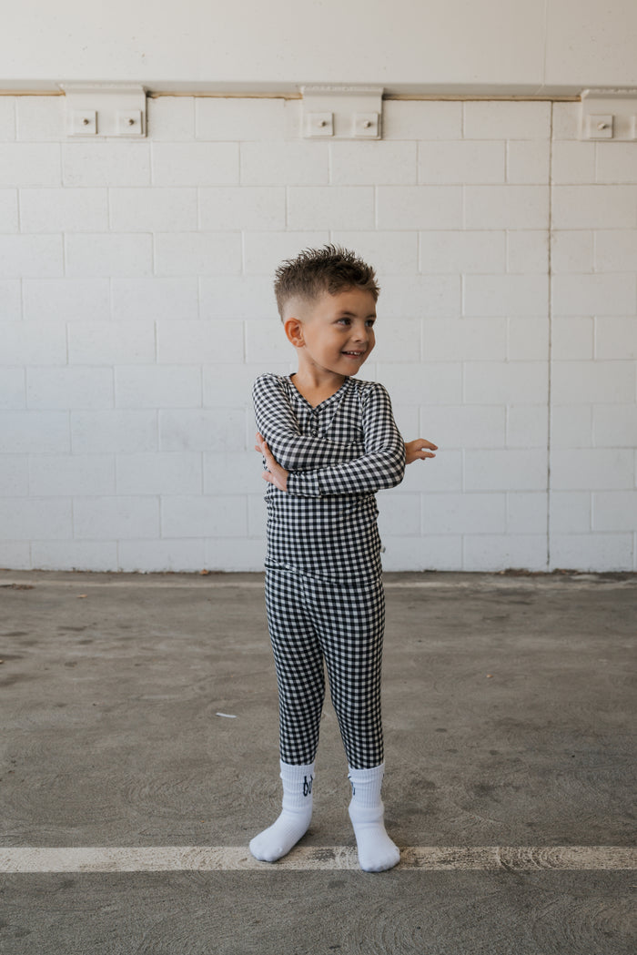A young child wearing forever french baby’s Bamboo Two Piece Pajamas in Black Gingham and white socks stands on a concrete floor, smiling and crossing their arms, with a white brick wall in the background.