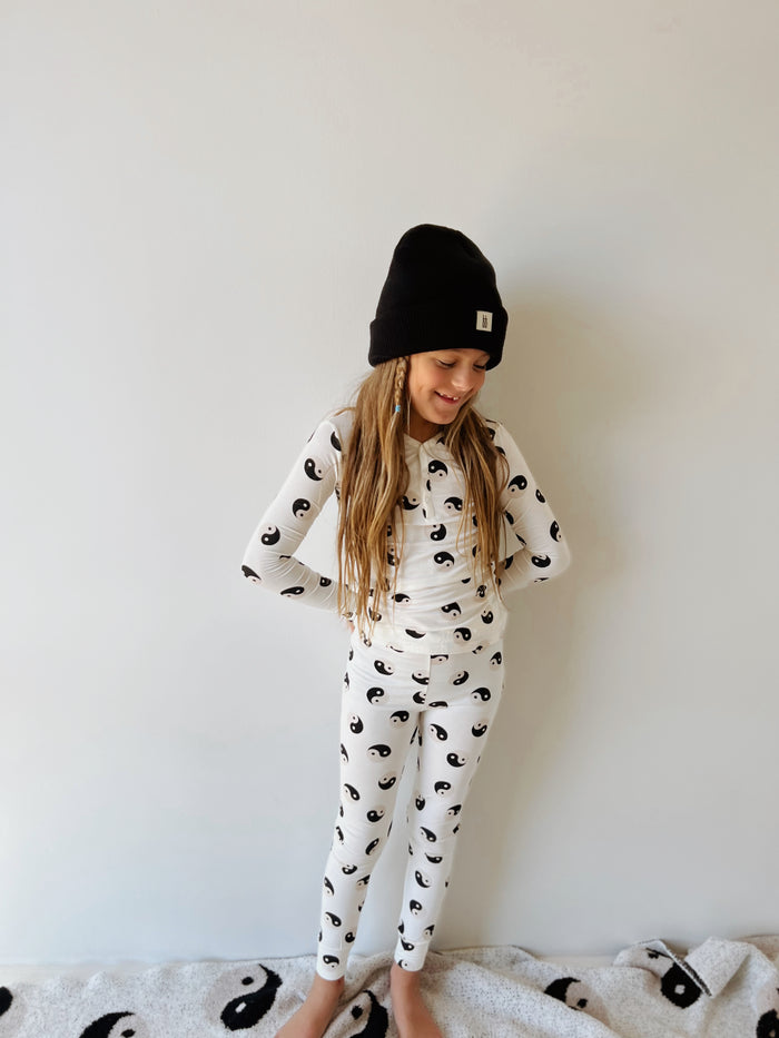 A young girl with long blonde hair stands against a plain wall, wearing a Bamboo Two Piece Pajamas | Black & White Yin Yang by forever French baby and a black beanie. She has a relaxed, cheerful expression with her hands resting on her hips. At her feet lies a blanket featuring a similar yin-yang design.