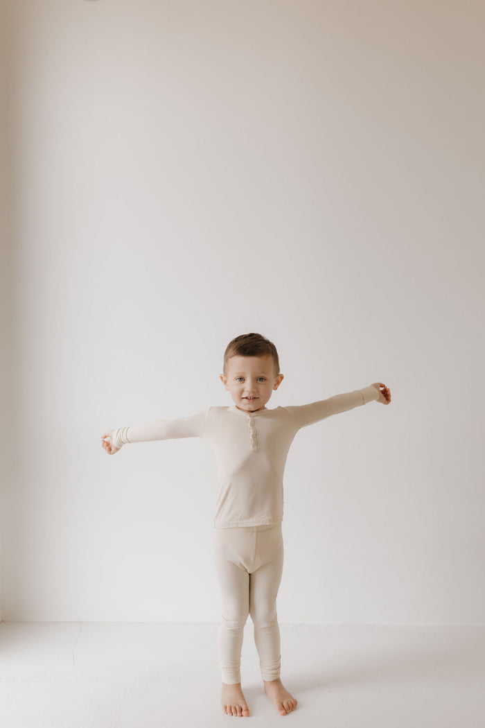 A young child stands barefoot in a minimalist room with arms outstretched, wearing the forever french baby Bamboo Two Piece Pajamas in Bone. Crafted from hypo-allergenic bamboo, the cozy beige pajamas contrast beautifully against the serene plain white background.