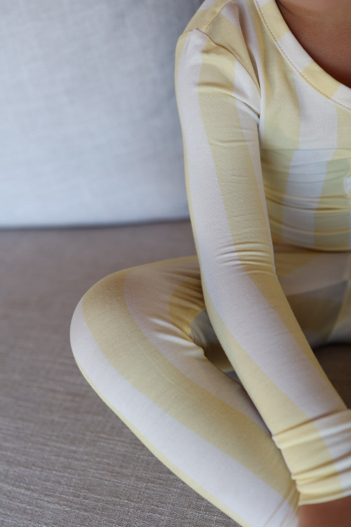 A child in forever french baby Bamboo Two Piece Pajamas | Butter (yellow and white stripes) sits on a light gray couch, with only the lower face and body shown.