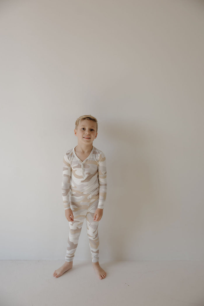 A young boy smiles gently, standing barefoot by a light wall in forever french baby's Bamboo Two Piece Pajamas | Camo 2.0—ideal for anyone seeking comfortable, durable sleepwear for kids.