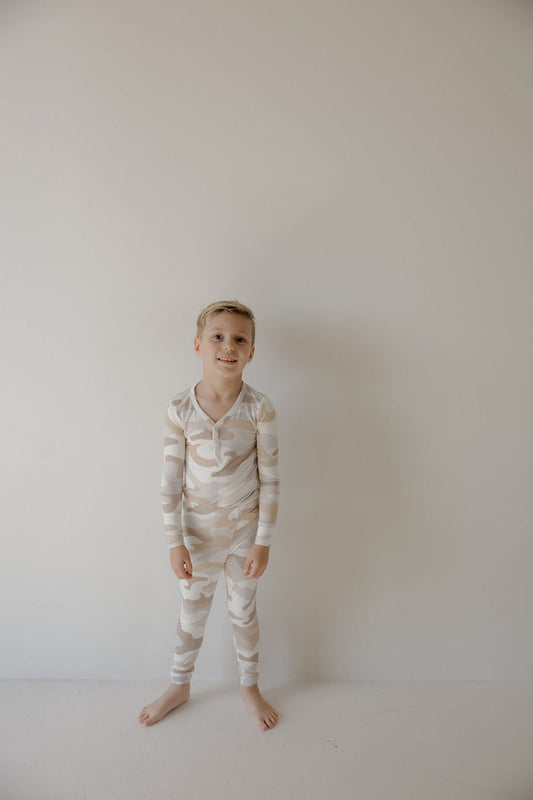A young boy smiles gently, standing barefoot by a light wall in forever french baby's Bamboo Two Piece Pajamas | Camo 2.0—ideal for anyone seeking comfortable, durable sleepwear for kids.