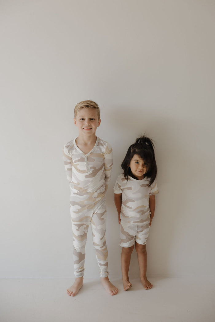 Two young children stand side by side in matching forever french baby Bamboo Two Piece Pajamas | Camo 2.0, made from hypo-allergenic bamboo for comfort—one smiles while the other has a neutral expression against a light wall.