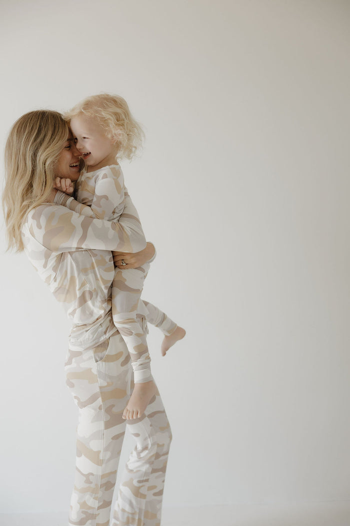 A smiling woman holds a young child as they wear matching forever french baby Bamboo Two Piece Pajamas in the Camo 2.0 print, standing against a plain light background.