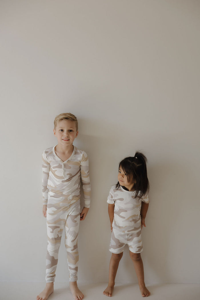 Two young children stand barefoot against a light wall. Both wear forever french baby Bamboo Two Piece Pajamas in Camo 2.0—the boy in long sleeves, the girl in short-sleeved hypo-allergenic matching clothing, gazing up at him.