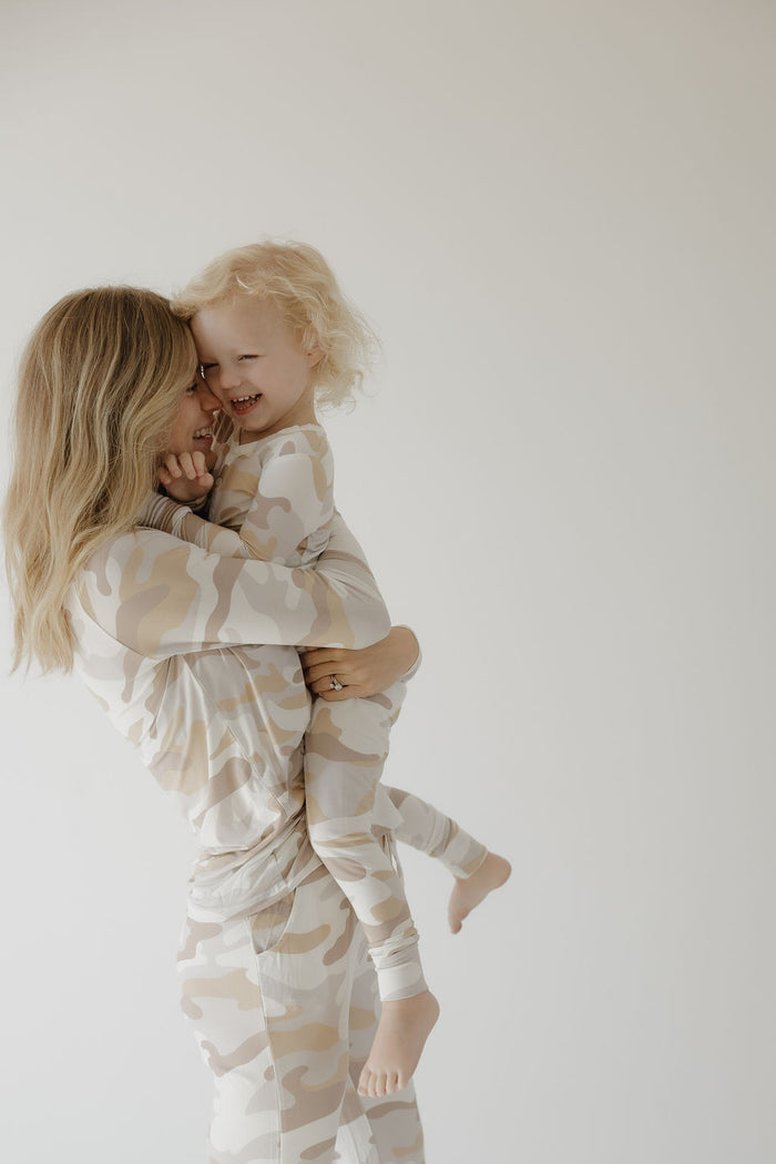 A woman holds a smiling child, both wearing matching forever french baby Bamboo Two Piece Pajamas | Camo 2.0, made from hypo-allergenic bamboo, against a light background, sharing a joyful moment.