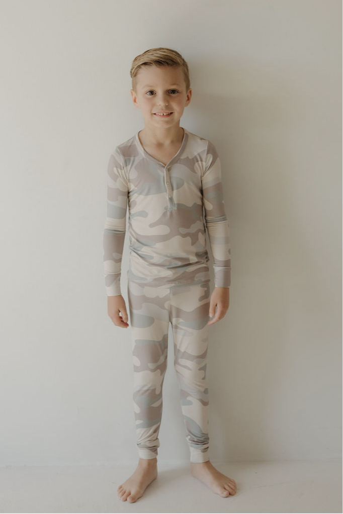 A young boy with short blond hair stands barefoot against a white wall, smiling in forever french baby's Bamboo Two Piece Pajamas | Camo—light-colored, long-sleeved bamboo pajamas made from breathable fabric.
