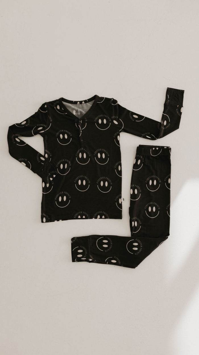 A relaxed set of Bamboo Two Piece Pajamas in Charcoal & White ff Smile from forever french baby is arranged on a light-colored surface. Made from breathable, hypo-allergenic fabric, the ensemble includes a top and matching pants, both adorned with the same whimsical white smiley face pattern.