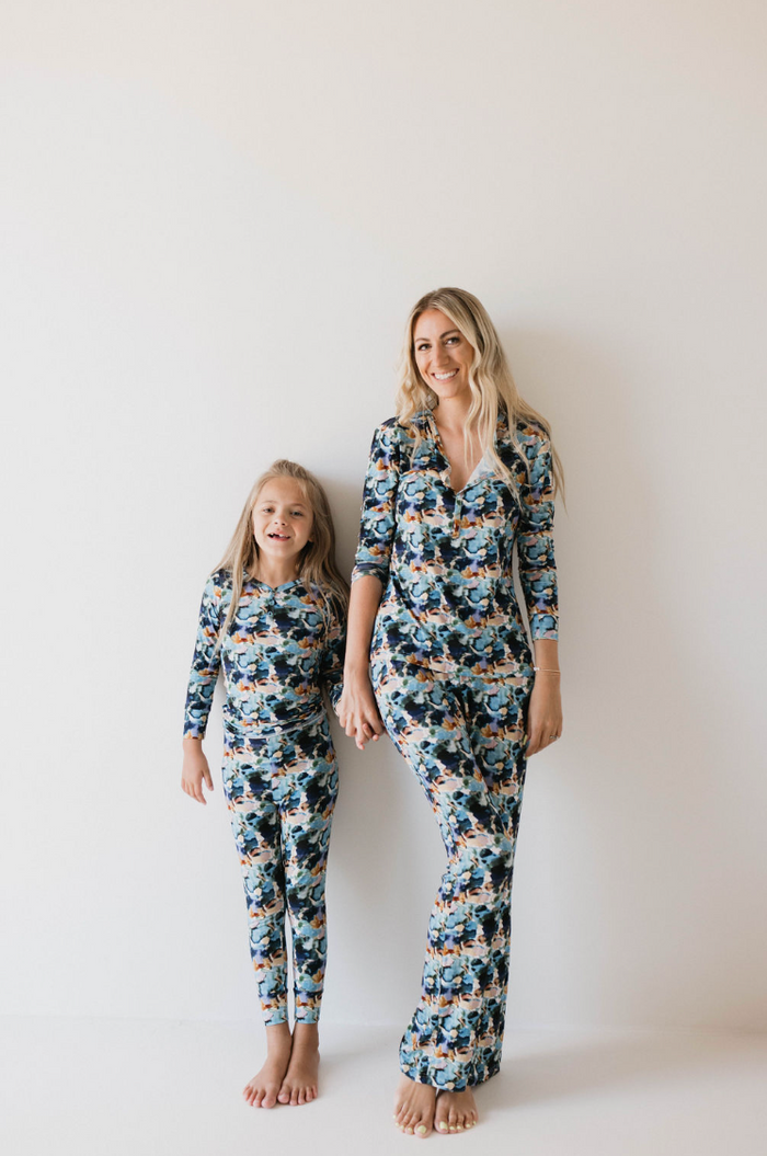 A woman and a young girl stand side by side against a plain white background, smiling and holding hands while wearing matching Bamboo Two Piece Pajamas in the Charli Print from forever french baby. The woman has long blonde hair, and the girl has her long blonde hair tied in a ponytail.