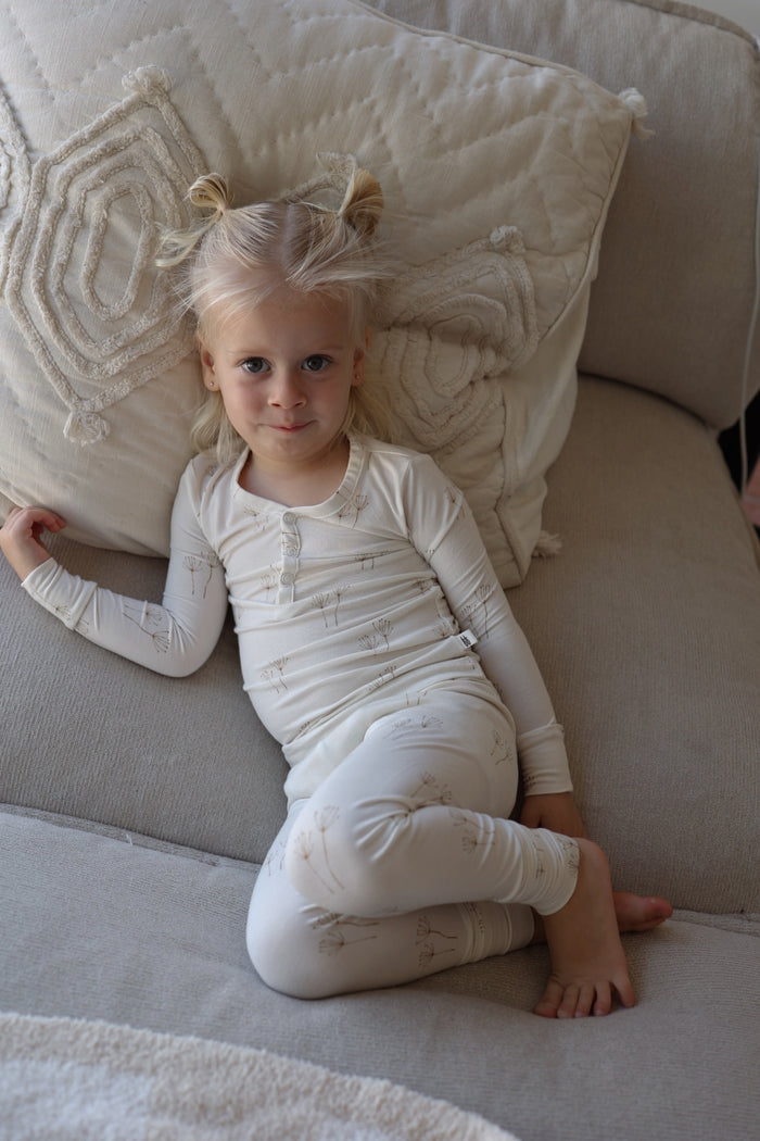 A young blonde child with pigtails, wearing forever french baby Bamboo Two Piece Pajamas in Dandelion, lounges on a beige couch with a textured pillow behind them and smiles gently at the camera.