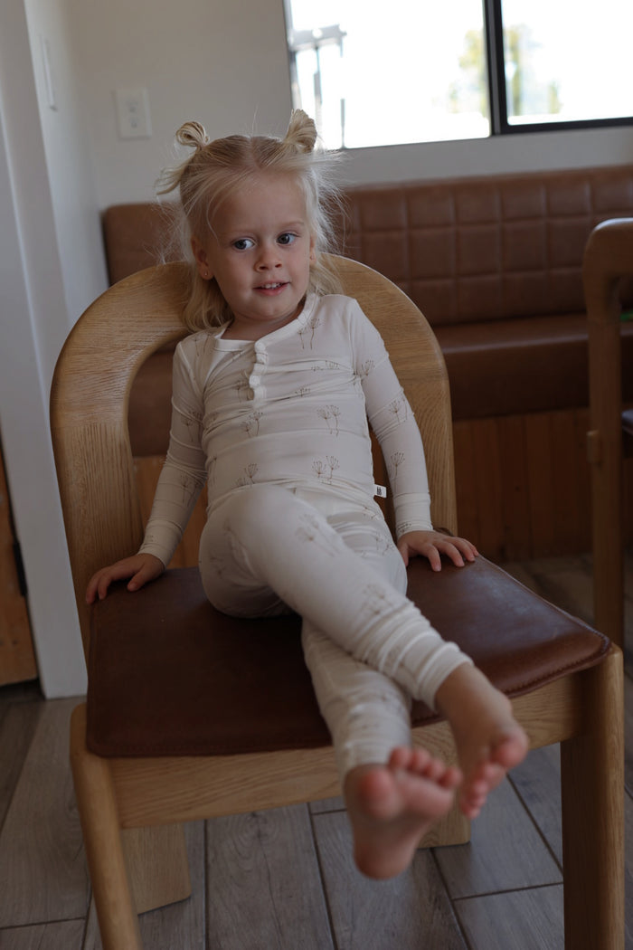 A young child with light hair in pigtails smiles while sitting on a wooden chair, wearing forever french baby’s Bamboo Two Piece Pajamas | Dandelion—soft, breathable kids sleepwear in a light color.