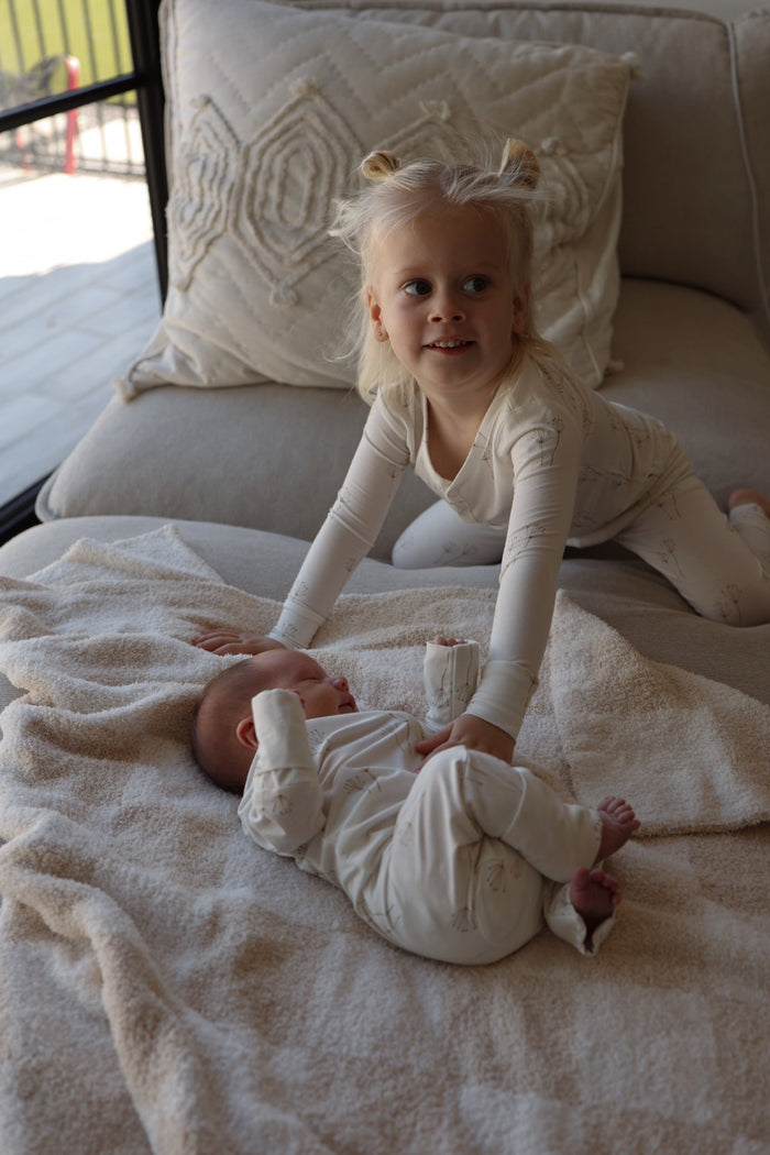 A blonde child in forever french baby’s Bamboo Two Piece Pajamas | Dandelion smiles while leaning over a baby on a blanket, both looking cozy in breathable bamboo sleepwear.