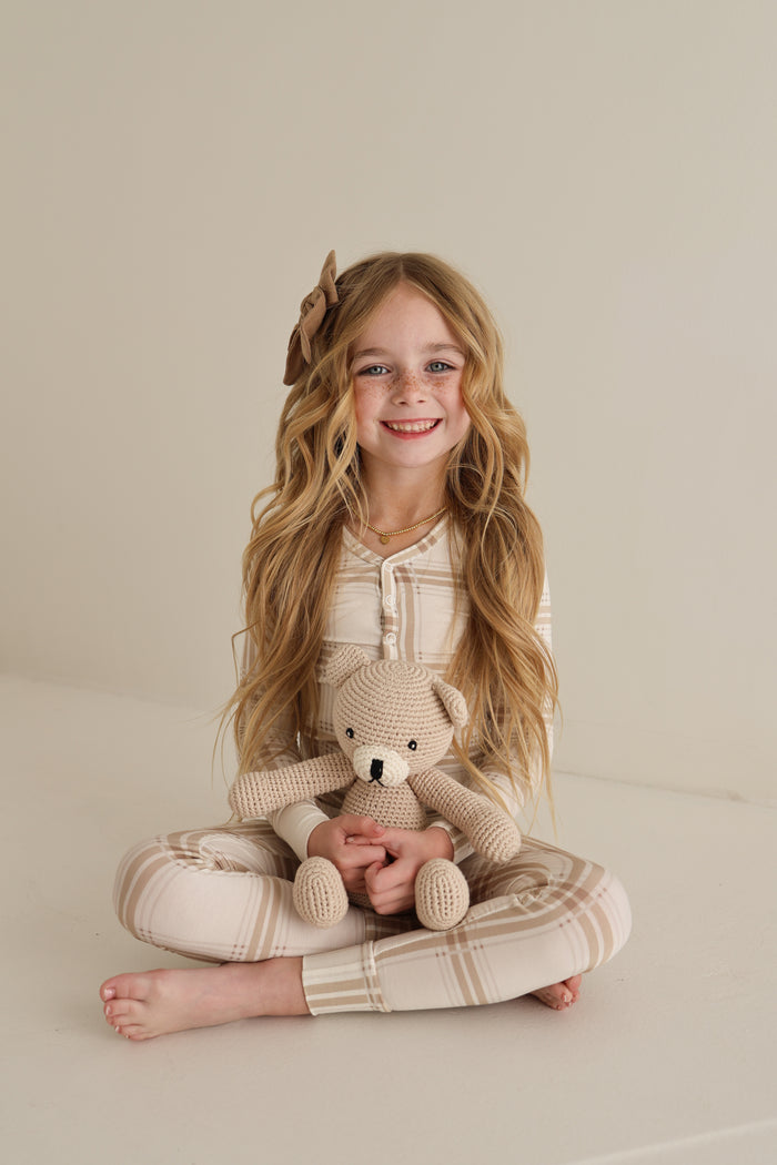 A young girl with long blonde hair and a brown bow sits cross-legged, smiling with a knitted teddy bear. She wears forever french baby Bamboo Two Piece Pajamas in Ellis Plaid—cozy and hypoallergenic for sensitive skin.