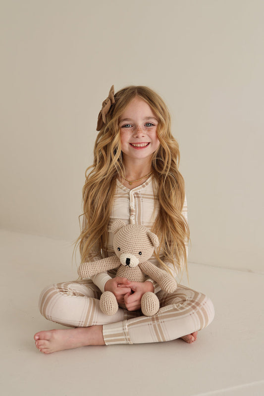 A young girl with long blonde hair and a brown bow sits cross-legged, smiling with a knitted teddy bear. She wears forever french baby Bamboo Two Piece Pajamas in Ellis Plaid—cozy and hypoallergenic for sensitive skin.