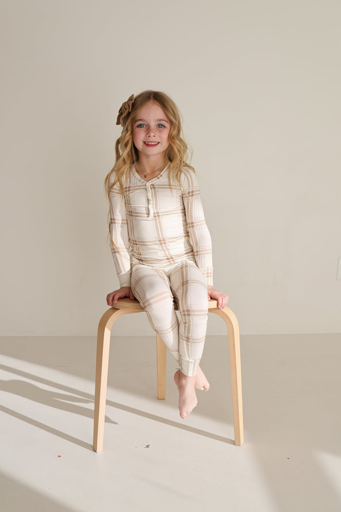 A young girl with long blonde hair, barefoot on a wooden stool, smiles at the camera in forever french baby’s Bamboo Two Piece Pajamas | Ellis Plaid—comfy kids sleepwear perfect for sensitive skin.
