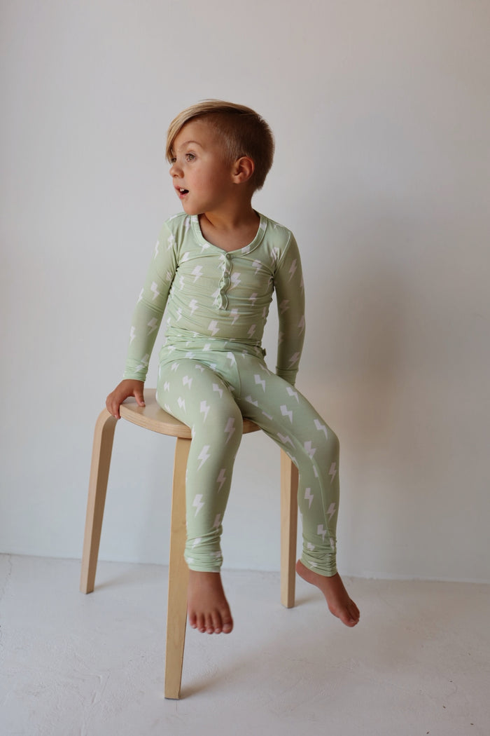 A young child with short blonde hair sits barefoot on a wooden stool, wearing forever french baby’s Bamboo Two Piece Pajamas | Flash—light green sleepwear with a white lightning bolt pattern—against a plain light background.