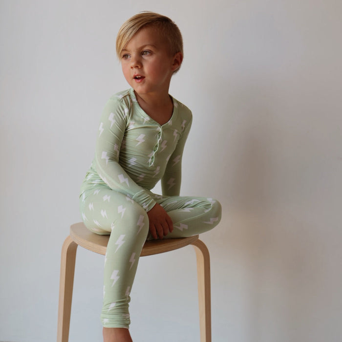 A young child with short blond hair sits cross-legged on a light wooden stool, wearing forever french baby's Bamboo Two Piece Pajamas | Flash in light green with a white lightning bolt pattern—perfect breathable sleepwear for all-night comfort.