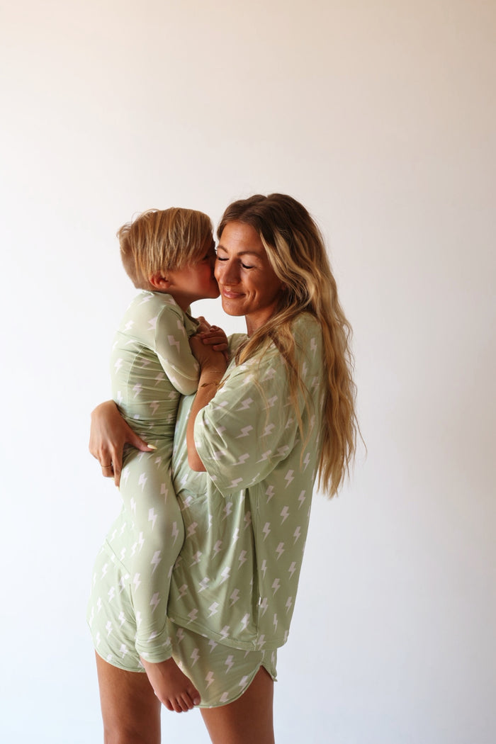A smiling woman holds a child who kisses her cheek as they wear matching forever french baby Bamboo Two Piece Pajamas | Flash—light green with white lightning bolts—against a plain background.